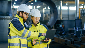 Two workers in hard hats and safety vests looking over data in a computer in a manufacturing warehouse